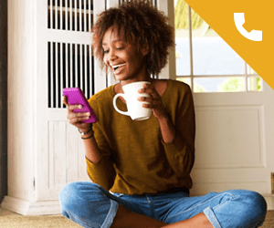 A young woman sitting on the floor looking at a smartphone and holding a mug